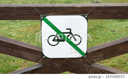 Clear and direct close-up shot of a metal prohibition sign indicating no cycling, mounted securely on a rustic brown wooden fence with a soft green grass background in a natural outdoor park setting Clear and direct close-up shot of a metal prohibition sign indicating no cycling, mounted securely on a rustic brown wooden fence with a soft green grass background in a natural outdoor park setting 143309557