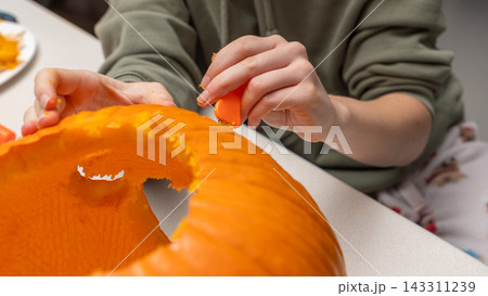 Person carving a pumpkin for Halloween, using a small tool to create intricate designs on the orange surface, with a table and pumpkin scraps visible in the background 143311239