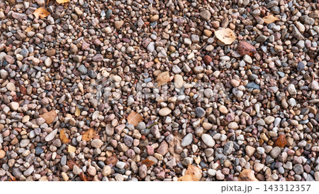 Dense pebble ground covered with mixed stones in earthy tones and scattered autumn leaves, creating natural outdoor texture for landscaping, background design, seasonal nature themes, and earthy. 143312357