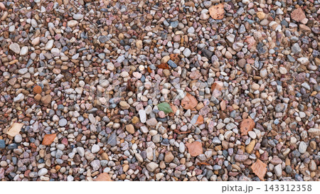 Multicolored small pebbles and gravel fill ground surface in dense natural pattern, with scattered dry leaves adding subtle seasonal detail, creating rough earthy texture for backgrounds and design. 143312358