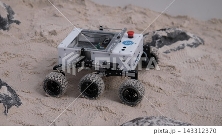 Compact wheeled rover crawls across soft sand among dark rocks, showing rugged experimental design, outdoor testing conditions, and exploration technology in harsh terrain Compact wheeled rover crawls across soft sand among dark rocks, showing rugged experimental design, outdoor testing conditions, and exploration technology in harsh terrain 143312370