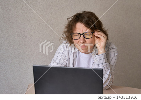 Woman Adjusting Glasses While Leaning Close To Laptop At Table In Plain Indoor Room With Beige Wall 143313916