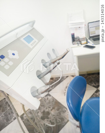 Dental Console With Digital Panel And Silver Handpieces Stands Beside Blue Chair, With Hanging 143314016