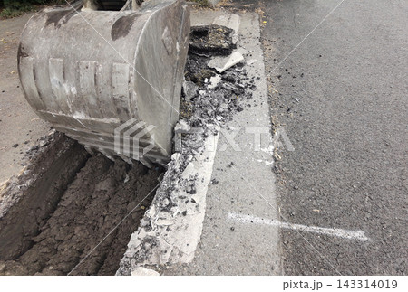 Steel Excavator Bucket Rests Over Narrow Trench Beside Broken Asphalt And Exposed Soil, With Loose Steel Excavator Bucket Rests Over Narrow Trench Beside Broken Asphalt And Exposed Soil, With Loose 143314019