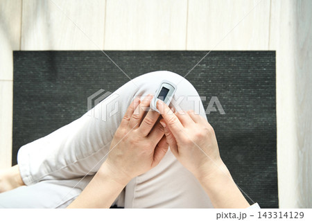 Person Operating Pulse Oximeter On Finger While Seated On Black Mat On Light Wooden Floor In Bright Person Operating Pulse Oximeter On Finger While Seated On Black Mat On Light Wooden Floor In Bright 143314129
