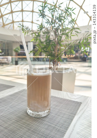 Tall Glass Of Pale Frothy Drink Stands With White Straw On Woven Placemat, Beside Potted Tree 143314139