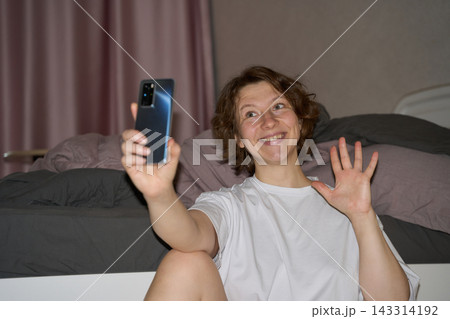Woman Waves Spread Fingers Beside Vertically Held Phone While Smiling Toward Screen, With Bent Knee Woman Waves Spread Fingers Beside Vertically Held Phone While Smiling Toward Screen, With Bent Knee 143314192