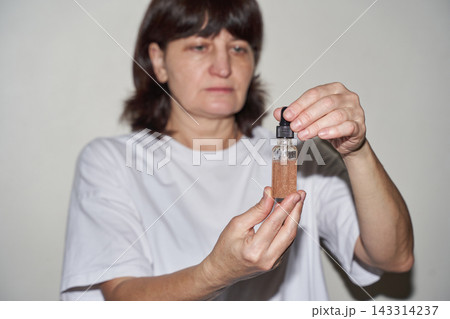 Middle-Aged Woman Examining Single Glass Dropper Bottle With Shimmer Serum And Removing Cap 143314237