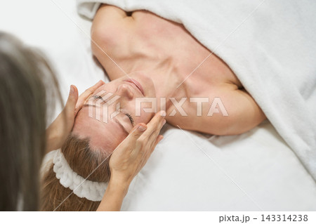 Woman Receiving Facial Massage With Closed Eyes And Headband On Spa Bed In Bright Treatment Room Woman Receiving Facial Massage With Closed Eyes And Headband On Spa Bed In Bright Treatment Room 143314238