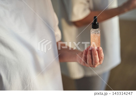 Woman Presenting Glass Dropper Bottle Of Glittering Serum In Soft Light Near Mirrored Companion Woman Presenting Glass Dropper Bottle Of Glittering Serum In Soft Light Near Mirrored Companion 143314247
