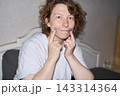 Woman Touching Blue Under Eye Patches With Both Index Fingers While Smiling At Camera In Bedroom 143314364