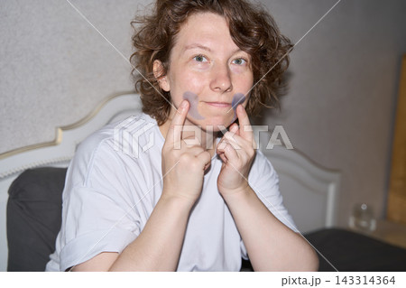 Woman Touching Blue Under Eye Patches With Both Index Fingers While Smiling At Camera In Bedroom Woman Touching Blue Under Eye Patches With Both Index Fingers While Smiling At Camera In Bedroom 143314364