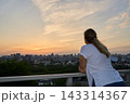 Young Woman Leaning On Balcony Railing Watching Distant City Skyline Under Warm Sunset Clouds 143314367