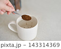 Spoon Releasing Instant Coffee Granules Into White Mug On Kitchen Counter With Hand Visible And Jar 143314369