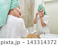 Woman Adjusting Under Eye Patches In Front Of Bathroom Mirror With Towel Wrapped Hair Standing 143314372