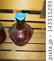 Textured glass carboy filled with dark red liquid for homemade wine fermentation. Blue latex glove airlock on wooden bench 143315293