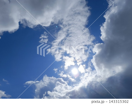 Bright blue sky with fluffy white and grey clouds. Intense sunlight shining through creating a glare Bright blue sky with fluffy white and grey clouds. Intense sunlight shining through creating a glare 143315302