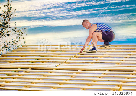 Measuring roof battens with tape measure while checking spacing and alignment, worker prepares wooden roof structure for installation of roofing materials. 143320974