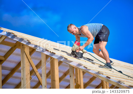 Securing timber battens over roofing underlay, worker uses nail gun while assembling wooden roof structure during construction. Securing timber battens over roofing underlay, worker uses nail gun while assembling wooden roof structure during construction. 143320975