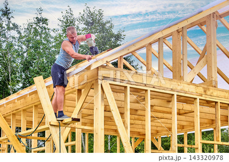 Carpenter fastens roof laths over weather barrier using compressed air nailer while standing on makeshift work platform. Carpenter fastens roof laths over weather barrier using compressed air nailer while standing on makeshift work platform. 143320978