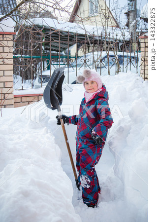 Young girl shoveling snow with large shovel in snowy yard near trees and fence in winter countryside Young girl shoveling snow with large shovel in snowy yard near trees and fence in winter countryside 143322875