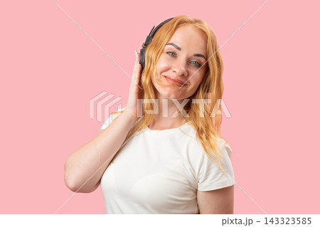 Young woman with red hair wearing wireless headphones, smiling while listening to audio content against a pink background, showcasing relaxation and happiness Young woman with red hair wearing wireless headphones, smiling while listening to audio content against a pink background, showcasing relaxation and happiness 143323585
