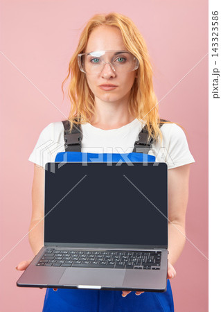 Young female worker wearing safety glasses and blue overalls standing against a pink background, holding a laptop with an empty black screen towards the viewer Young female worker wearing safety glasses and blue overalls standing against a pink background, holding a laptop with an empty black screen towards the viewer 143323586