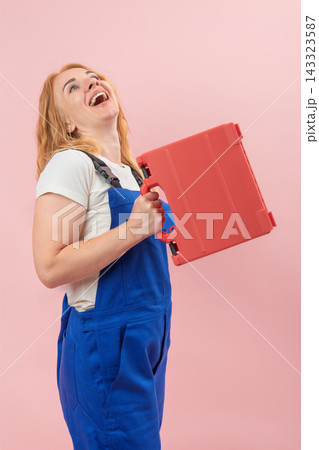 Cheerful young woman in blue overalls smiling widely and looking up, holding a red toolbox, embodying success and professional happiness against a pink background Cheerful young woman in blue overalls smiling widely and looking up, holding a red toolbox, embodying success and professional happiness against a pink background 143323587