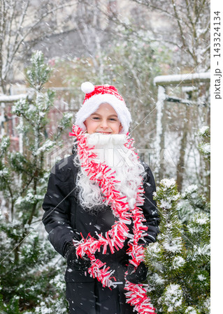 Child dressed as Santa with white beard red tinsel garland standing outdoors in snowy winter garden Child dressed as Santa with white beard red tinsel garland standing outdoors in snowy winter garden 143324134
