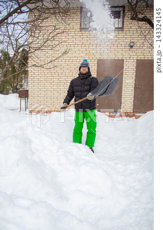 Man in winter jacket shoveling snow with shovel in front of brick house after heavy snowfall 143324145