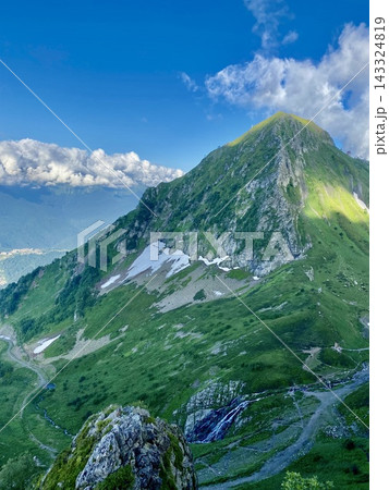 Green alpine mountain rises above melting snow and cascading summer waterfall. Green alpine mountain rises above melting snow and cascading summer waterfall. 143324819