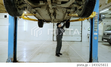 Professional car mechanic inspecting the chassis of a lifted vehicle, performing maintenance and repairs in a modern automotive repair shop 143325811
