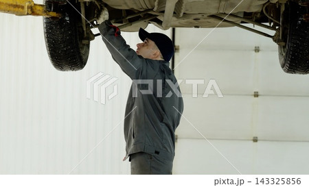 Skilled mechanic examining undercarriage components beneath lifted vehicle, wearing uniform and cap in brightly illuminated automotive workshop 143325856