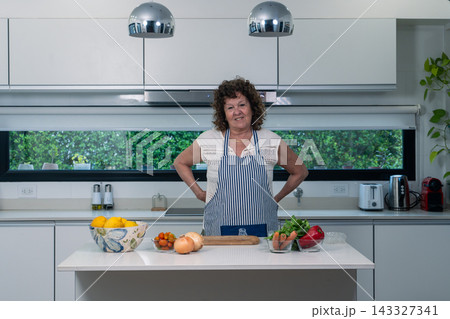 Old female chef posing proudly in a sleek modern kitchen with fresh ingredients arranged before 143327341