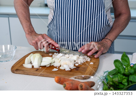 Senior woman methodically dicing white onions on a wooden board in a sunlit kitchen Senior woman methodically dicing white onions on a wooden board in a sunlit kitchen 143327353
