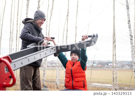 Father helping his son with street pull-down machine exercise to train back muscles and arms in autumn park, family support concept 143329313
