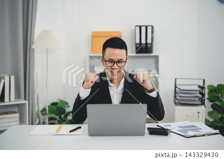 Excited businessman celebrating success at modern office desk with laptop and documents, smiling man wearing glasses and suit Excited businessman celebrating success at modern office desk with laptop and documents, smiling man wearing glasses and suit 143329430