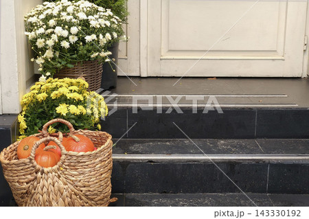 Autumn entrance decorated with chrysanthemums and pumpkins in a woven basket, creating a cozy seasonal scene with rustic charm and Halloween atmosphere. 143330192