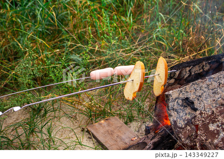 Man prepares bread and sausages on skewers sitting by campfire in nature. Outdoor activities, camping and tourism concept 143349227