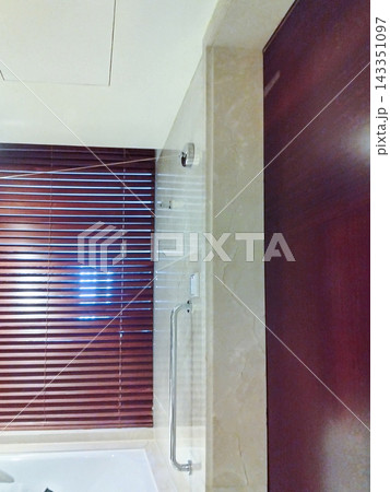 Bathroom interior with glass shower partition, chrome grab bar, wall fixtures, wooden window blinds, and dark wooden door in a residential washroom Bathroom interior with glass shower partition, chrome grab bar, wall fixtures, wooden window blinds, and dark wooden door in a residential washroom 143351097