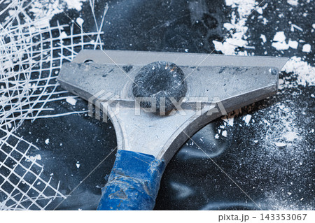 Close up of a metal scraper lying on a dark surface surrounded by plaster debris and torn mesh 143353067