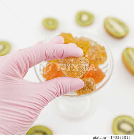 Hand in pink glove selecting colorful fruit gummies surrounded by fresh kiwi slices Hand in pink glove selecting colorful fruit gummies surrounded by fresh kiwi slices 143355013
