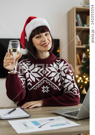 A Caucasian woman wearing a festive Santa hat and a snowflake sweater raises a glass of white wine while working at her desk during the holiday season A Caucasian woman wearing a festive Santa hat and a snowflake sweater raises a glass of white wine while working at her desk during the holiday season 143355554