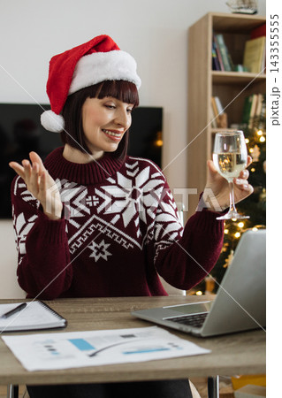 A smiling Caucasian woman wearing a Santa hat and festive sweater raises a glass of wine while video conferencing on her laptop during the holidays 143355555