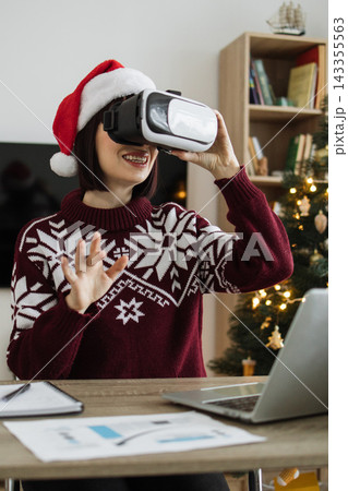 A Caucasian woman wearing a Santa hat and festive sweater uses a virtual reality headset while sitting at a desk with a laptop and Christmas tree in the background 143355563