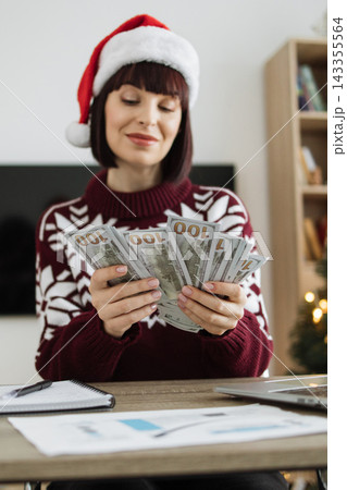 A Caucasian woman wearing a festive sweater and Santa hat counts a fan of US dollar bills at a desk with a laptop and papers A Caucasian woman wearing a festive sweater and Santa hat counts a fan of US dollar bills at a desk with a laptop and papers 143355564