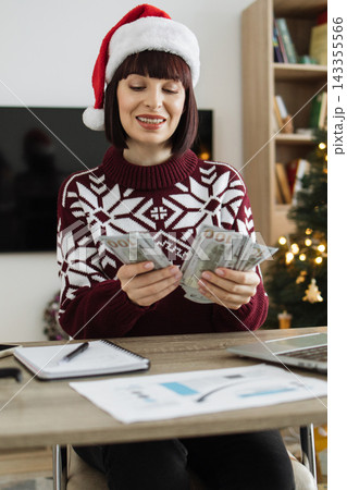 A woman wearing a Santa hat and festive sweater counts US dollar bills while sitting at a desk with a laptop and papers 143355566