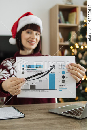A smiling woman wearing a Santa hat points to a bar graph showing positive growth trends during the holiday season A smiling woman wearing a Santa hat points to a bar graph showing positive growth trends during the holiday season 143355610