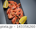 On a black stone board on a gray background are frozen shrimps, framed on both sides by lemon slices. Concept: symmetrical citrus frame, enhancing the bright emphasis on seafood. High quality photo 143356260