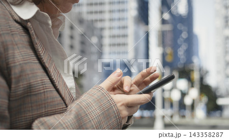 Businesswoman using smartphone, close up against city background during daylight. Female professional checks mobile device outdoors with metropolitan street softly blurred Businesswoman using smartphone, close up against city background during daylight. Female professional checks mobile device outdoors with metropolitan street softly blurred 143358287